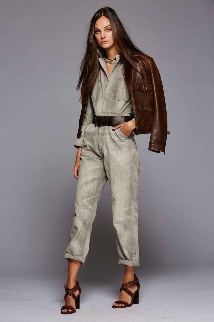 a full shot of a female model wearing a light grey denim jumpsuit with a brown leather jacket draped over her shoulders. she accessorizes with a brown belt, brown sandals, and a silver necklace. her long brown hair is loose, and she is posing against a grey background in a studio setting. the overall style is casual yet chic and elegant.