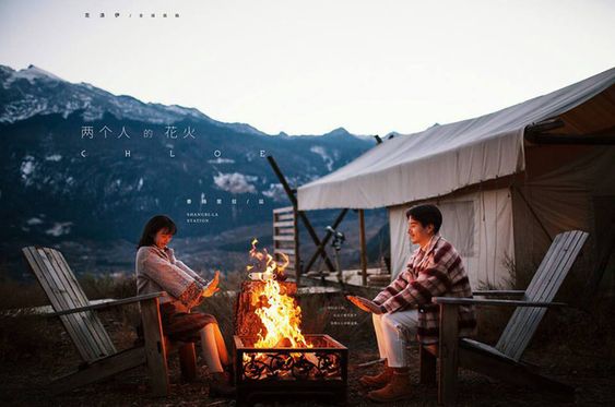 a couple is sitting in front of a campfire in a snowy mountain setting. the man is wearing a plaid shirt and jeans, and the woman is wearing a grey jacket and jeans. they are both smiling and looking at the fire. there is a tent in the background, and the sun is setting behind the mountains.