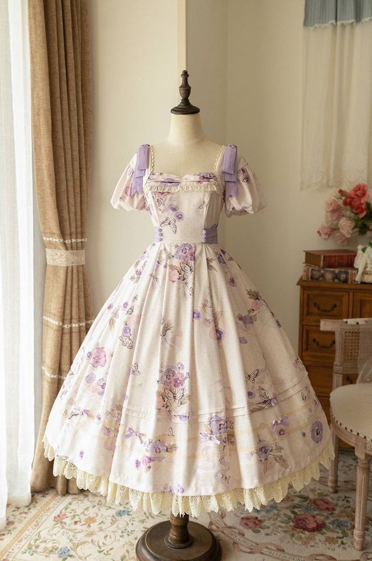 a fulllength view of a creamcolored lolita fashion dress with a floral print in shades of purple. the dress is displayed on a mannequin in a welllit room. the dress has a square neckline, puff sleeves, and a full skirt. the skirt is decorated with lace trim. the dress is styled with a white petticoat. the background of the image shows a window with curtains, a wooden dresser, and a rug.