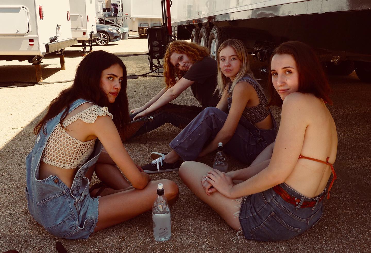 the image shows four young women sitting on the ground outdoors, in what appears to be the setting of a film or television production.  several trailers are visible in the background. the girls are casually dressed in denim overalls, a crochet top, and a bikini top.  they appear relaxed and friendly, enjoying a break on a sunny day. the overall aesthetic has a slightly vintage or 70s vibe.  two water bottles are visible in the foreground.