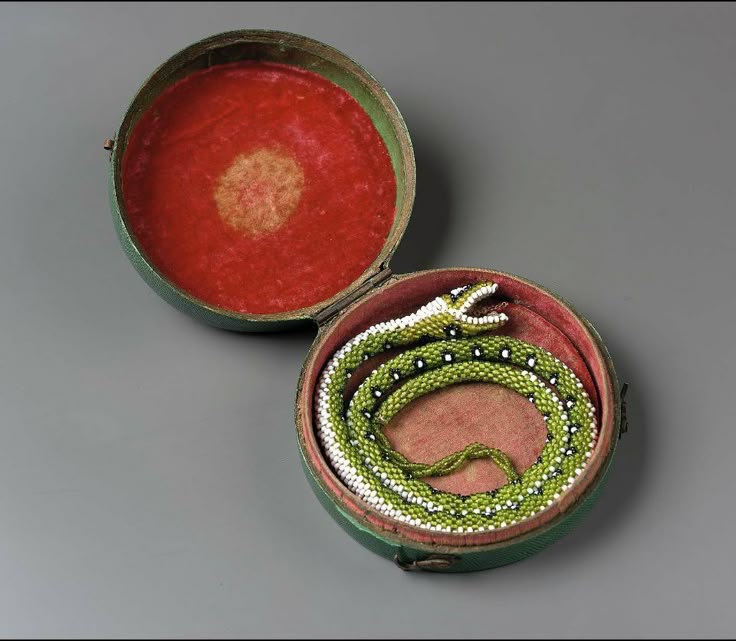 a beaded snake coiled inside a round box with a red velvet lining. the snake is primarily green with white accents. the box is green with a red velvet lining. the box lid is open and also lined with red velvet. the snake's head is raised with its mouth open. the snake is coiled on top of a red velvet cushion.