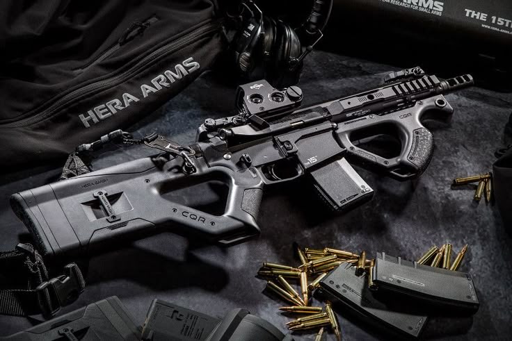 a black hera arms cqr rifle is laid out on a dark surface alongside ammunition and magazines. there are also accessories such as a bag, headset, and box with text about small arms. the rifle features a unique design with a cqr stock and foregrip. a red dot optic is mounted on the upper receiver. cartridges and magazines are scattered near the rifle.