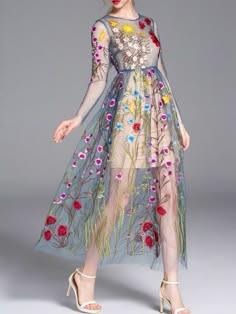 a woman wearing a beautiful floral embroidered sheer dress with long sleeves and a flowing skirt, standing on a gray background. she is wearing white heels and is looking straight ahead. the dress is very detailed with a floral pattern throughout.