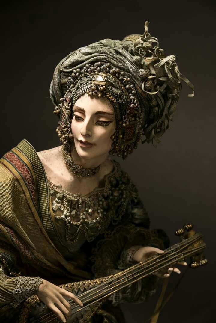 a mannequin or doll of a woman in antique clothing plays a lute. she wears a large, decorative turban headpiece adorned with beads and a triangular jewel. she has dark eye makeup and an elaborate dress with pearllike beads around the neckline and embroidered detailing. she holds the lute with her left hand touching the strings.