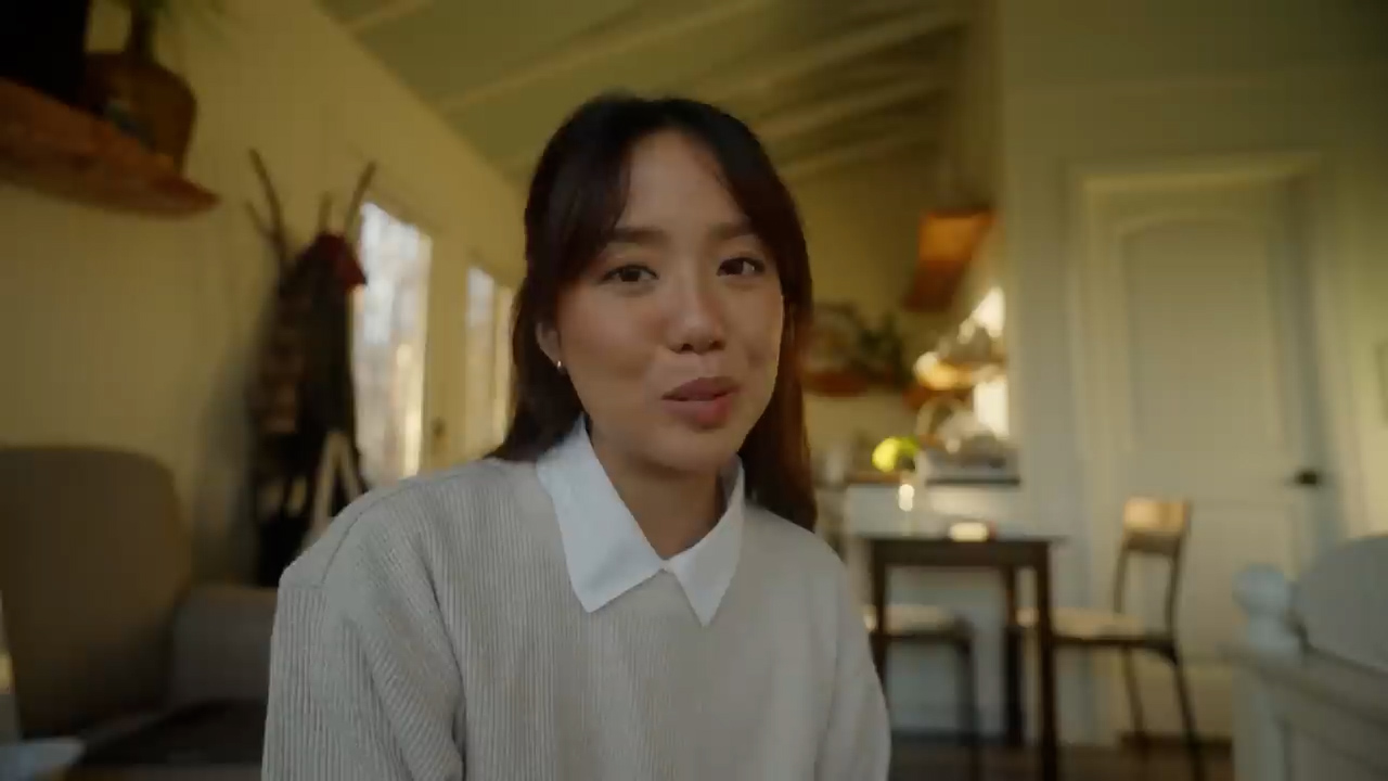 a young asian woman with shoulderlength dark hair is sitting in a cozy living room and talking to the camera. she's wearing a beige sweater with a white collar. the room is bright and airy, with warm natural light coming from the windows. the overall feel of the image is inviting and relaxed.