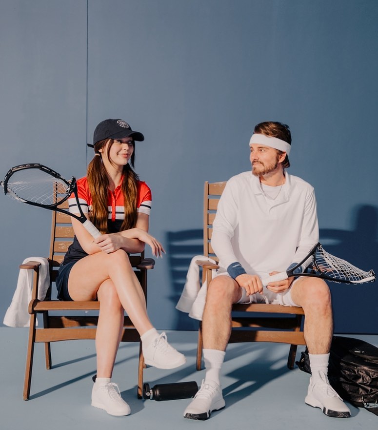 a woman and a man are sitting on chairs in a tennis setting. the woman is wearing a navy and red tennis outfit with a baseball cap, white tennis shoes, and is holding a tennis racket. the man is wearing a white tennis outfit, a white headband, white socks and tennis shoes, and is also holding a tennis racket. they are sitting next to each other with a blue background. a white towel is draped over the back of each chair, and a black water bottle is on the floor near the woman's chair. a tennis bag is on the floor near the man's chair.