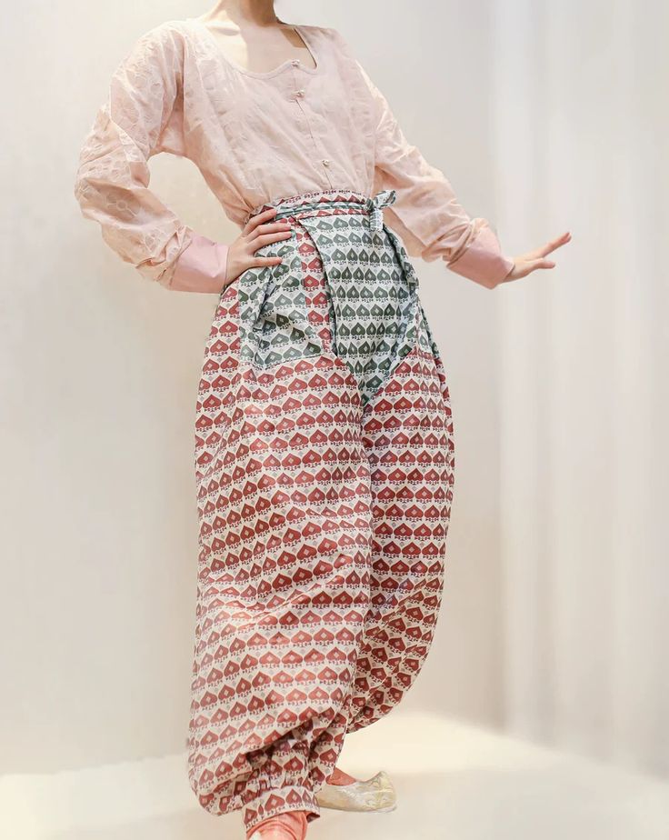 a woman is standing in front of a white wall wearing a pink blouse and patterned pants. the blouse is a light pink color and has long sleeves. the pants are a widelegged style with a red and green pattern. she is posing with one hand on her hip and the other outstretched.