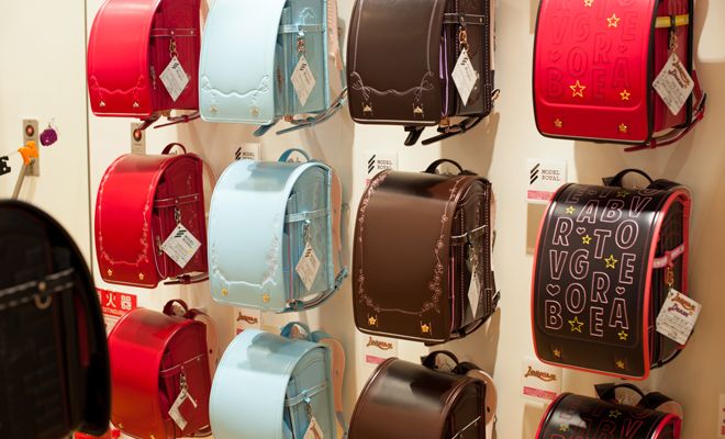 an image of a wall display featuring multiple japanese randoseru backpacks in various colors including red, blue, brown, and black. the backpacks are made of leather and are hanging on the wall. they are likely for sale in a retail store.