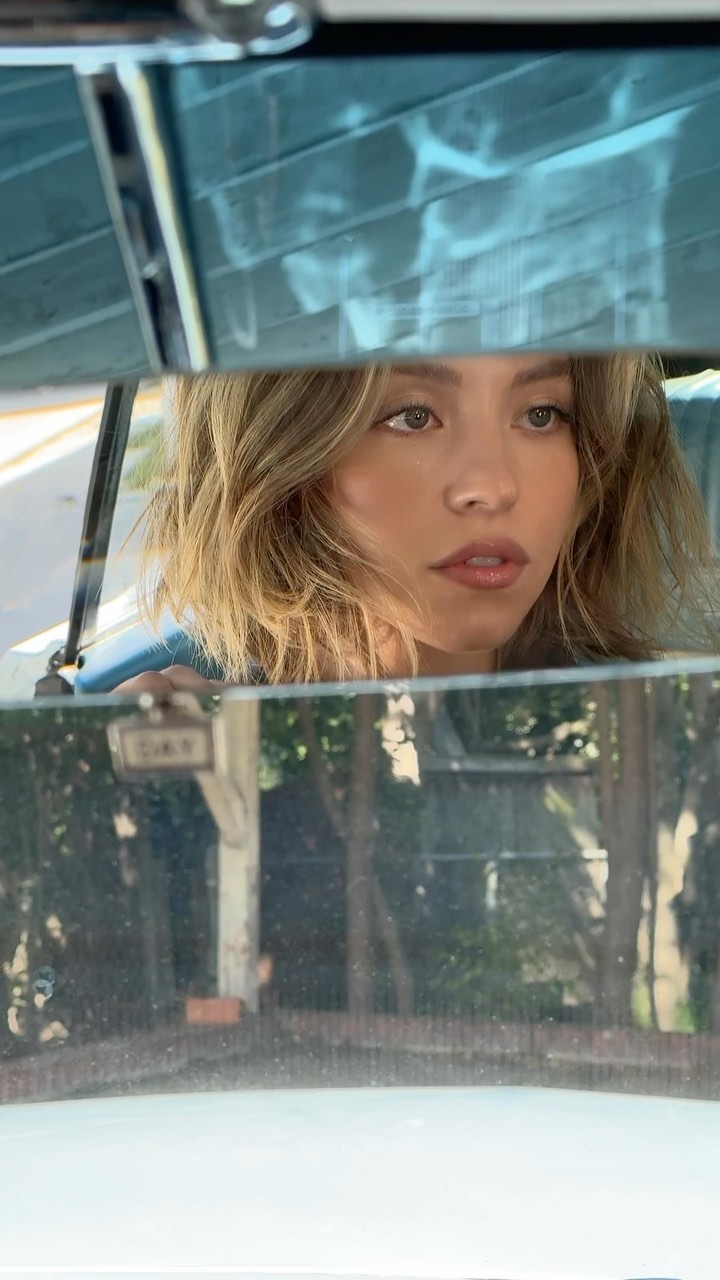 a closeup shot of a young woman with shoulderlength blonde hair sitting in a car. the image is taken from the inside of the vehicle, looking out through the windshield. the woman's face is partially obscured by reflections in the car's interior, giving a dreamy, atmospheric feel.  she appears pensive or thoughtful, looking off to the side. the reflections on the interior show a hint of trees and a blurry background. the overall mood is serene and contemplative.
