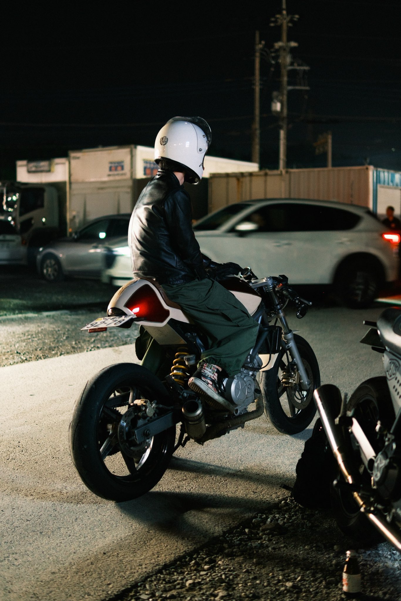 a rider in a white helmet is sitting on a custom motorcycle on a city street at night. the rider is wearing a black leather jacket and dark pants.  the motorcycle has a white and red paint job with a dark exhaust pipe. the rider is looking ahead at the road. there is a white car in the background, and the street is lit by the headlights of the cars.
