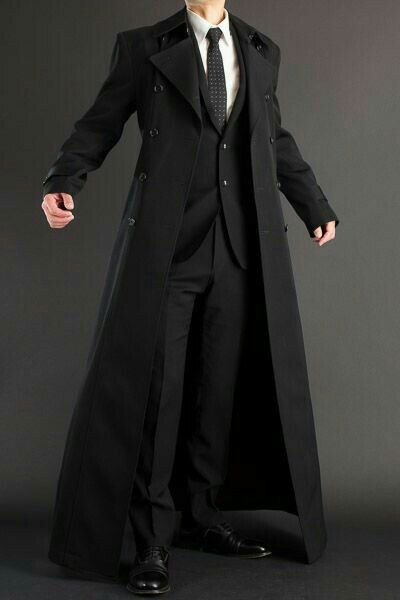 a man in a black long coat and matching black suit stands against a gray backdrop, he has a thin black tie,  his pose is casual with his hands in his pockets.
