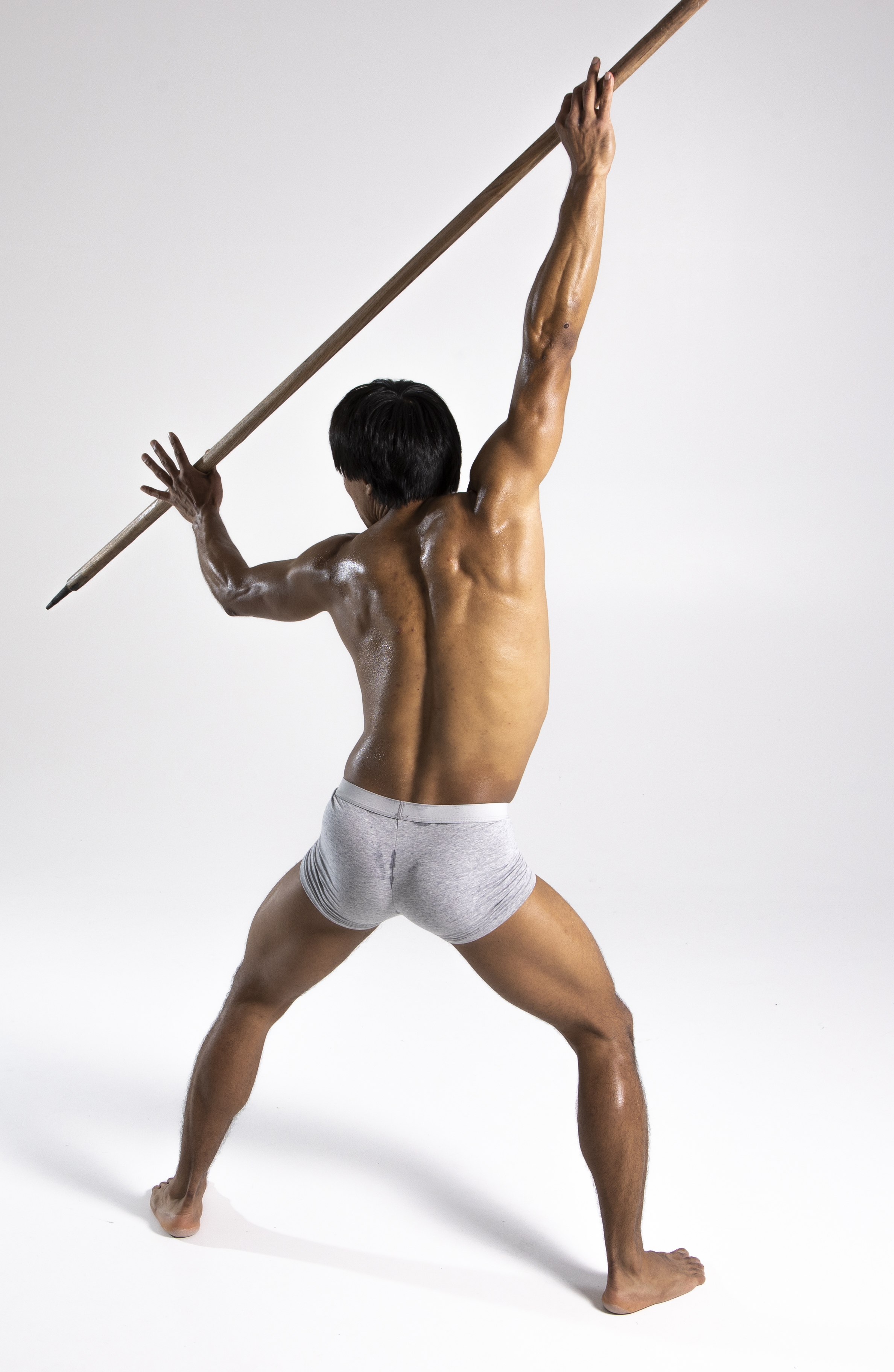 a muscular man stands with his back to the camera, holding a spear above his head. he is barefoot and wearing underwear. the background is white.