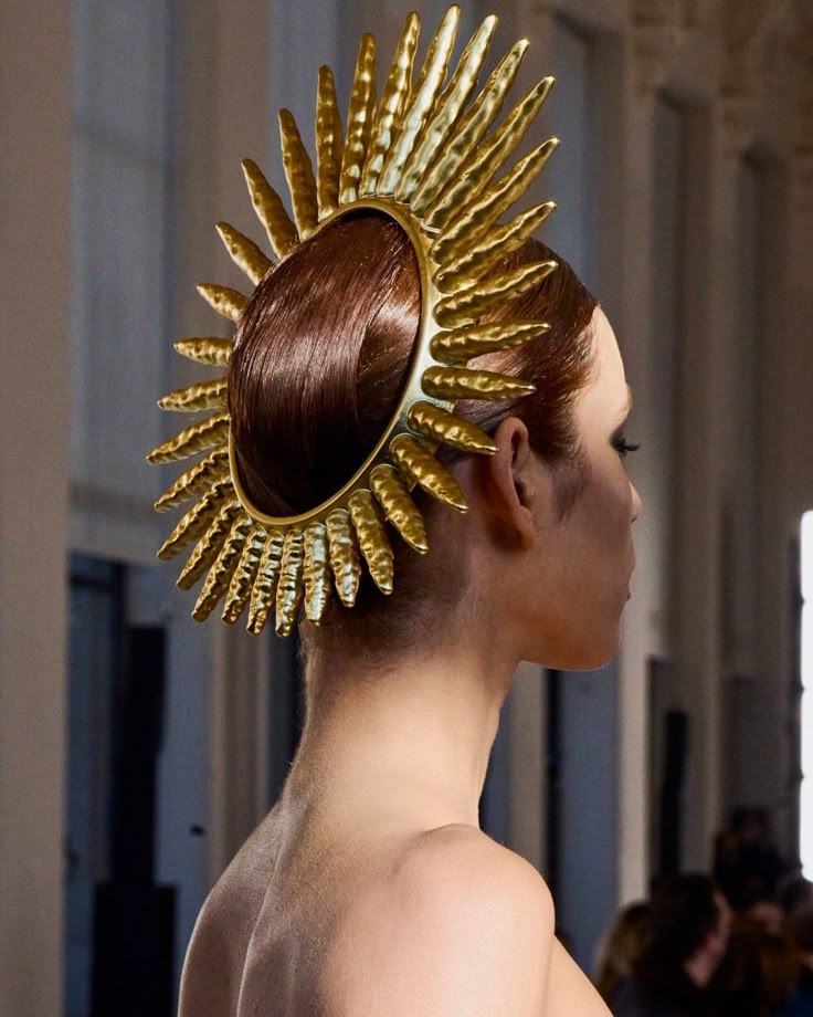 a woman with her back to the viewer is modeling a golden headdress. the headdress consists of a golden ring with spikes protruding from it. her hair is styled up into the center of the ring. she is wearing a sleeveless top that exposes her shoulders. the background is blurred and features a stage.