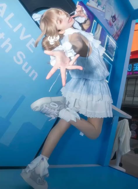 a young blonde woman in a blue dress is wearing white sneakers and smiling, she is leaning up against a blue wall while her arms are stretched out and she's making a hand gesture. the background of the image is a blurry cityscape.