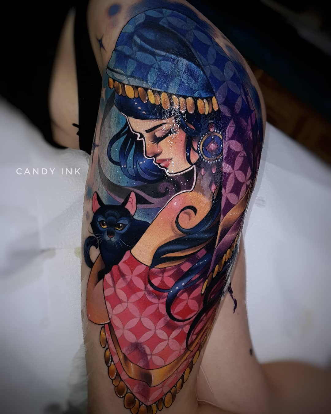 a tattoo on someone's upper thigh of a woman in a blue headscarf holding a black cat. the woman is wearing a pink patterned dress. the background of the tattoo is blue and purple.