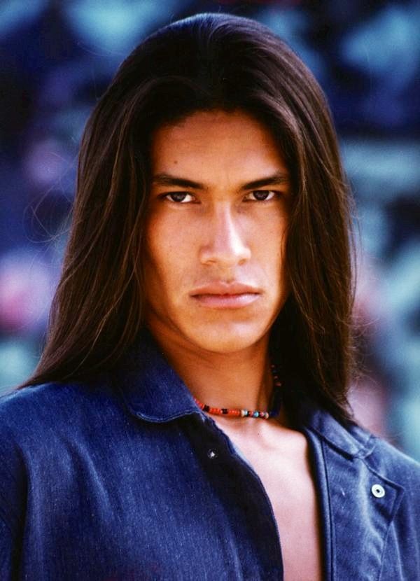 a portrait of a native american man with long, flowing brown hair. he has a serious expression and is wearing a denim shirt and a beaded necklace. the background is an outdoor setting with blurred foliage.