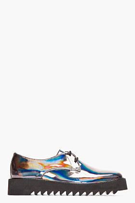 a pair of women's iridescent silver oxfords with a chunky, serrated platform sole. the shoes have a laceup closure and a metallic, holographic finish that shimmers with different colors in the light.  they are presented on a white background.