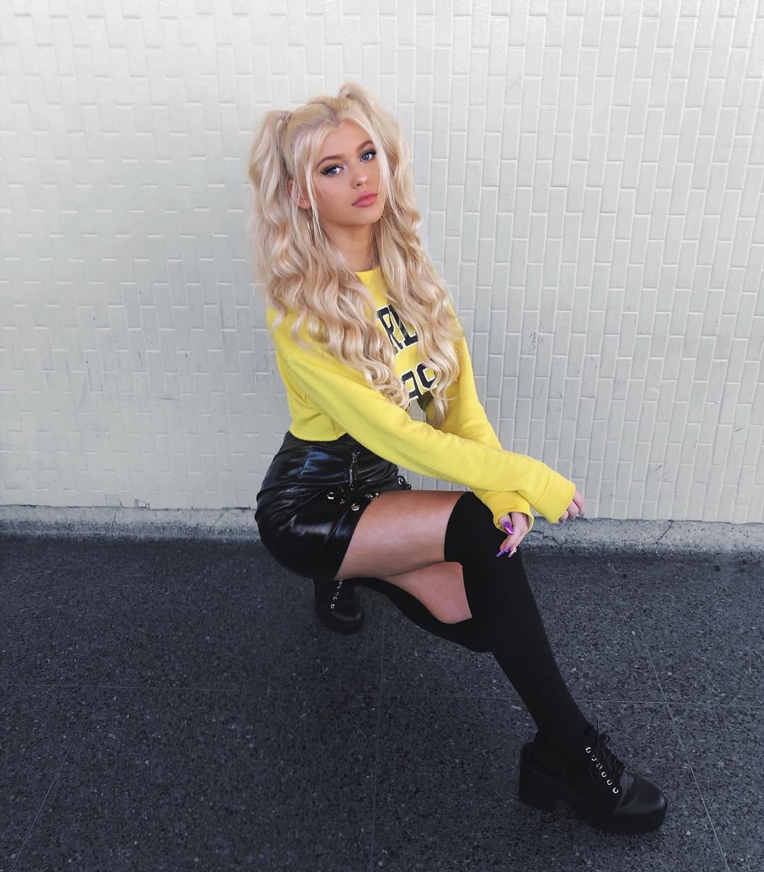 a woman with long blonde hair in pigtails is crouching against a white brick wall. she is wearing a yellow sweater, a black skirt, black socks, and black shoes. the pavement is visible in the foreground.