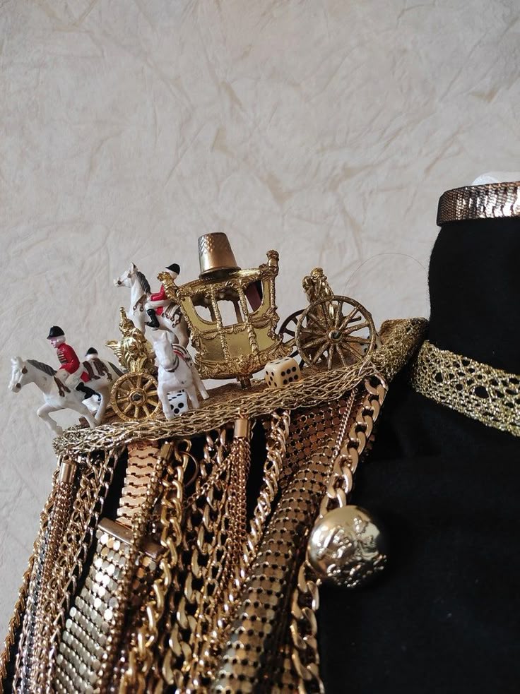 the image shows a mannequin decorated with gold jewelry and miniature figurines. the mannequin is black, and adorned with a variety of gold chains, a gold choker, and a gold band. on top of the mannequin there are toy figures including miniature horses, riders dressed in red, and a gold carriage with a thimble on top. there are also dice placed among the figurines. the arrangement appears decorative and elaborate.