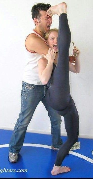 a man and a woman are practicing martial arts in a studio. the woman is holding her leg up in the air, and the man is holding her foot with his hand. they are both looking at each other. the woman is wearing black pants, and the man is wearing jeans. the floor is blue and white, and the walls are white. they are both wearing white belts. the man has a white shirt on and the woman has a white shirt on. the woman has brown hair and the man has brown hair. the woman is barefoot and the man is wearing shoes. they are both looking focused. they are both smiling. they are both having fun.
