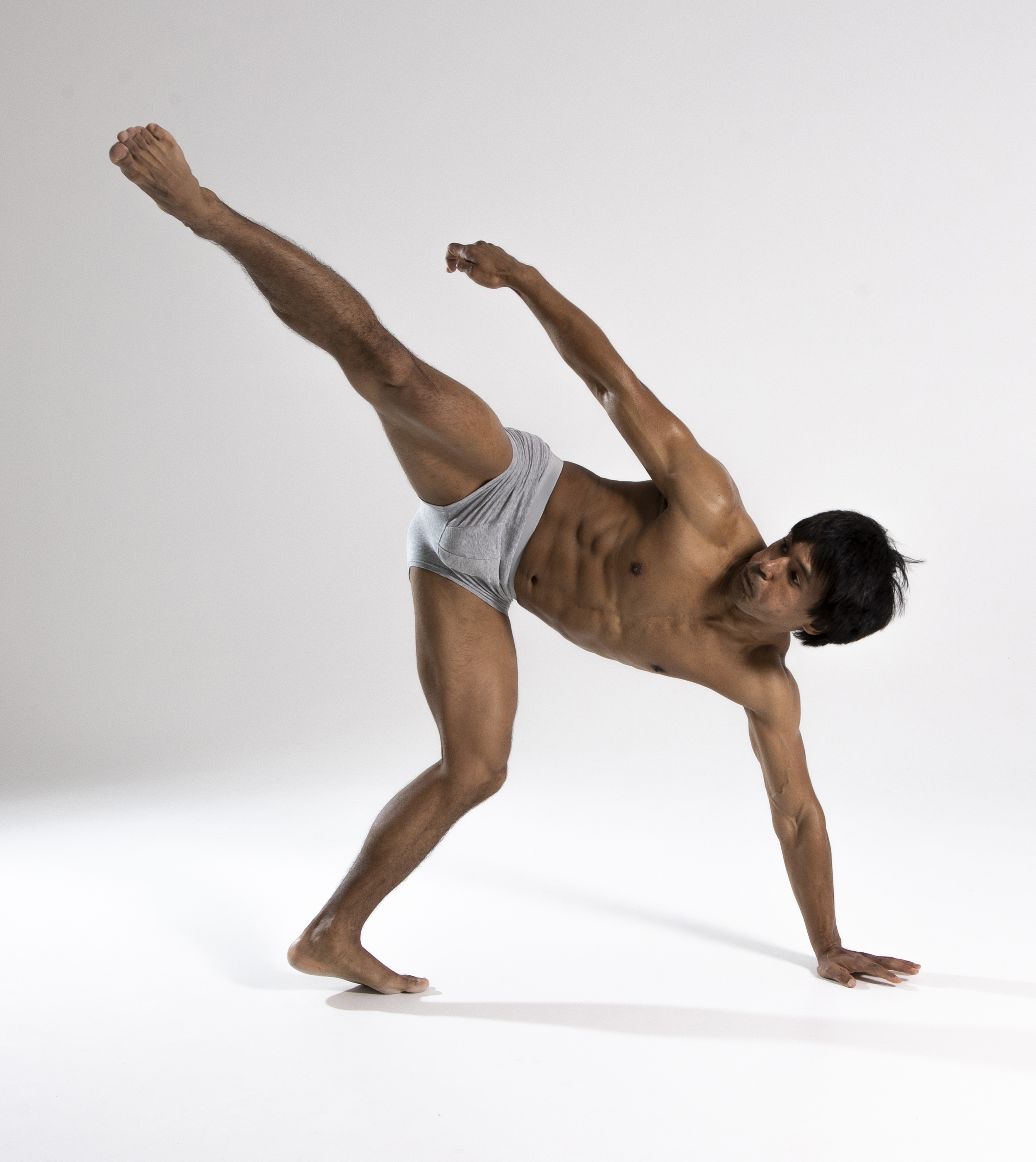 a shirtless man wearing gray underwear is captured in a dynamic dance pose against a plain white background. he is barefoot, with one leg extended high in the air to the left and his body angled forward and to the right. his right hand is on the floor, providing balance, and his gaze is directed off to the side. the man has dark hair and an athletic build with visible abs. the lighting emphasizes the contours of his body, highlighting his musculature.