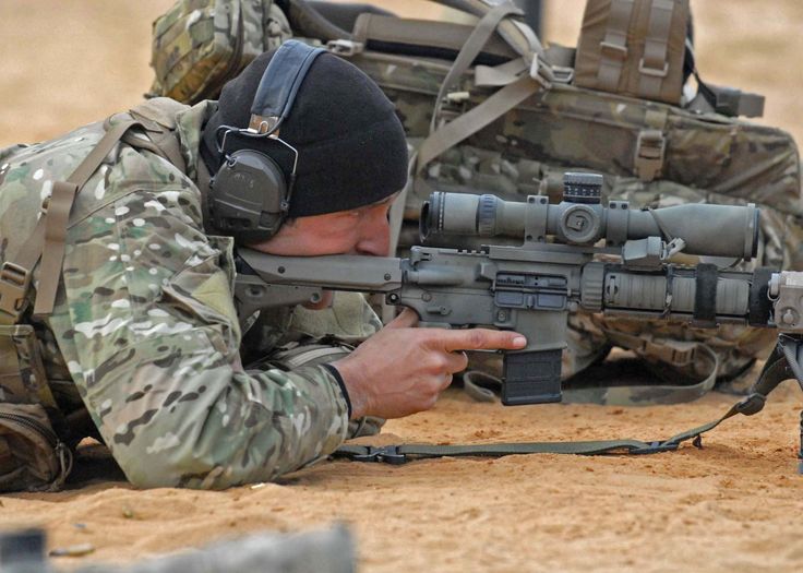 a soldier in camouflage gear is lying prone on the ground, aiming a scoped rifle. the soldier is wearing a black beanie hat and ear protection. the rifle has a bipod and is resting on a sandbag. the background is blurry, with a sandcolored environment.  the soldier's focus is intense as he aims down the scope, highlighting the precision and discipline required for this type of operation.