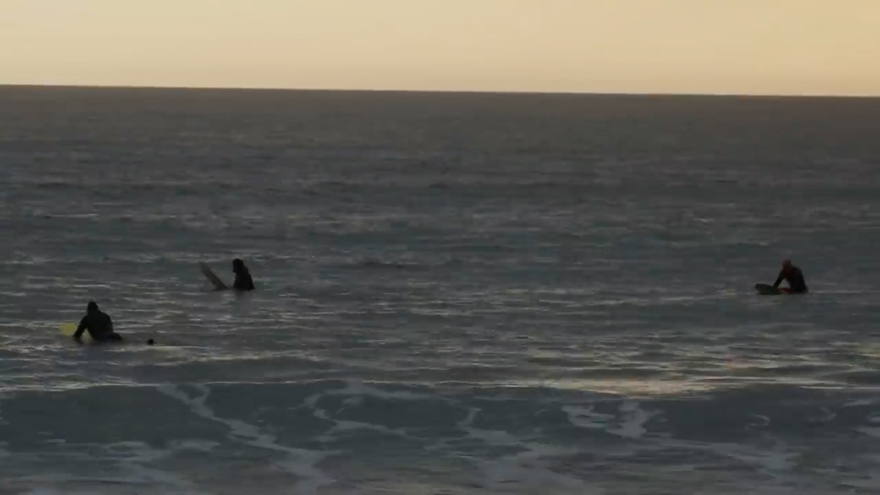 three surfers wait for a wave on their surfboards. they are spread out in the ocean and silhouetted by the setting sun. the lighting is dim, making the water and sky appear in dark shades of blue and gray.