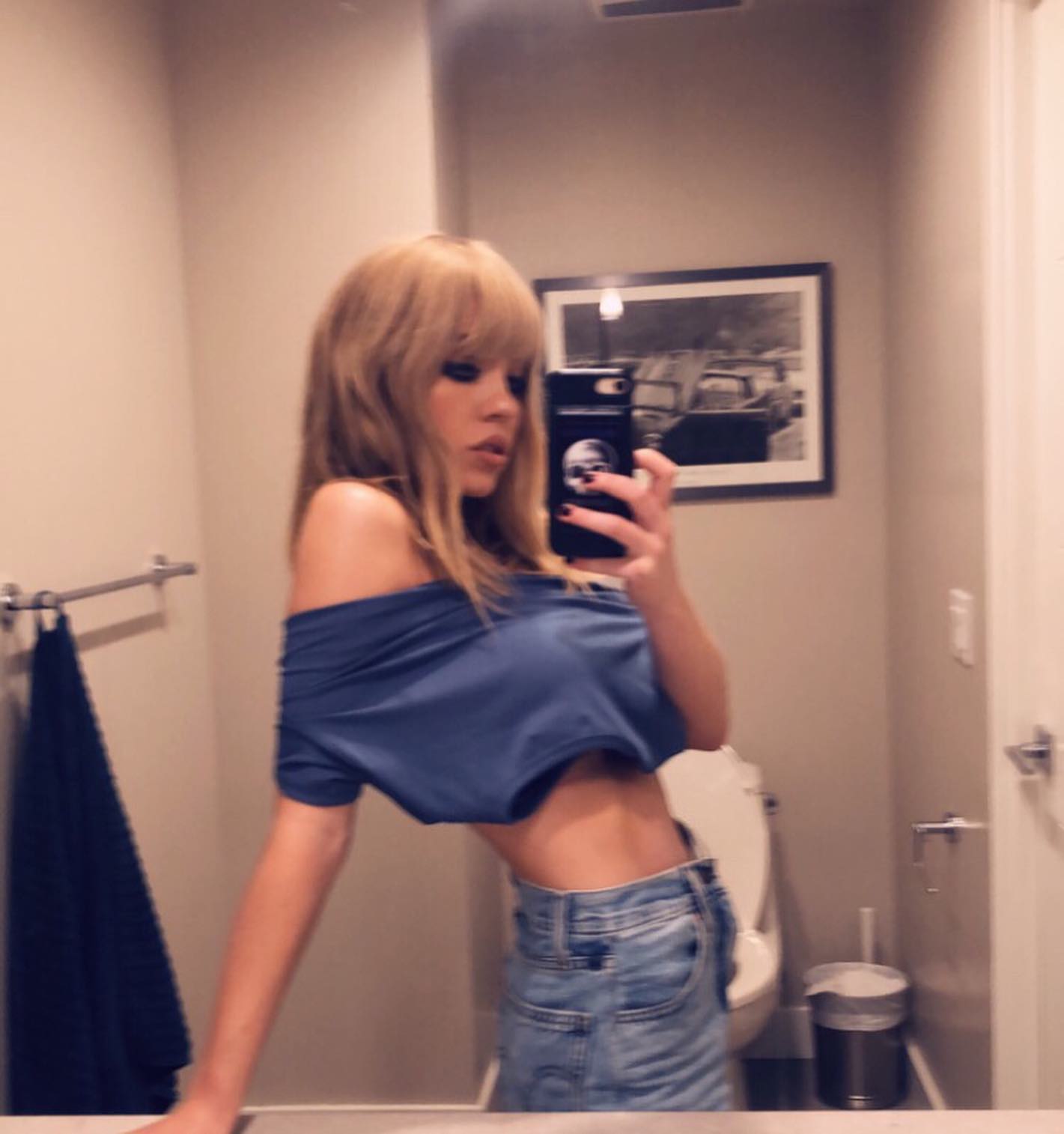 the image is a selfie taken by a young blonde woman in a bathroom. she's wearing a blue offtheshoulder cropped top and light wash highwaisted jeans.  her hair is styled with blunt bangs. she's holding a smartphone with a skulldesign phone case, taking a picture of herself in a mirror. the bathroom has gray walls and a toilet is visible in the background. the overall style is casual and the selfie is taken from a slightly angled perspective, showcasing her body.