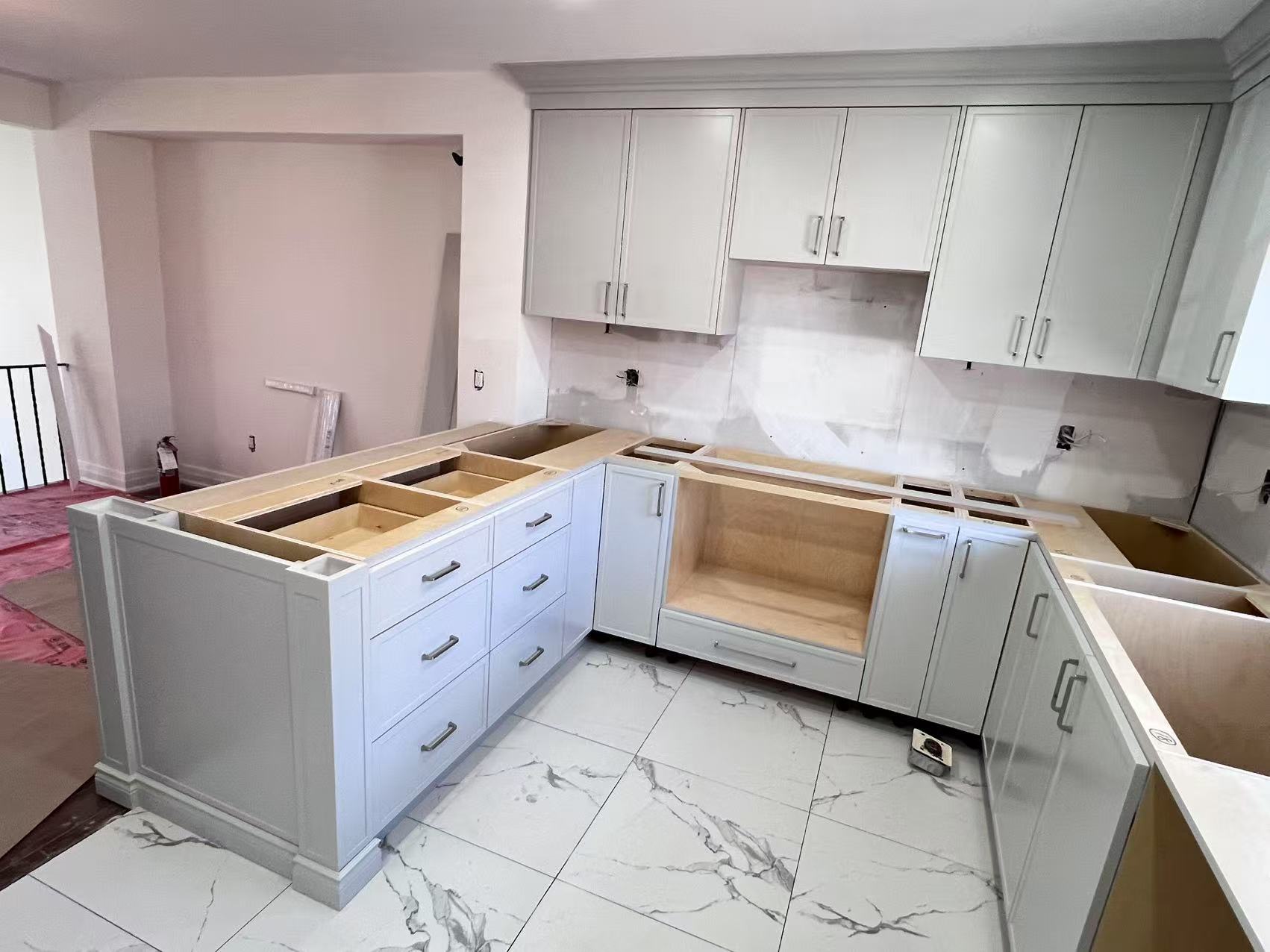 an interior shot depicts an underconstruction kitchen. predominantly white cabinets are installed along the wall and as part of a kitchen island, which features several drawers. the countertops are awaiting finishing touches. the floor is covered with marble tiles. the kitchen is a work in progress, representing a remodeling project or new construction.