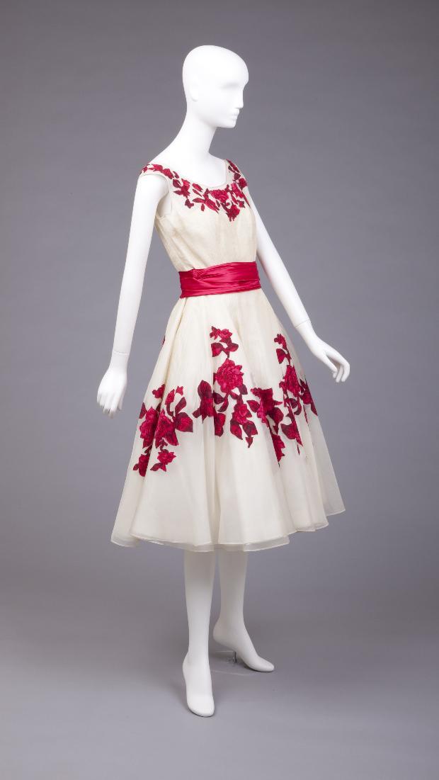 a white 1950s party dress with red floral embroidery displayed on a mannequin in a studio setting. the dress features a fitted bodice, a full skirt, and a red satin ribbon at the waist.