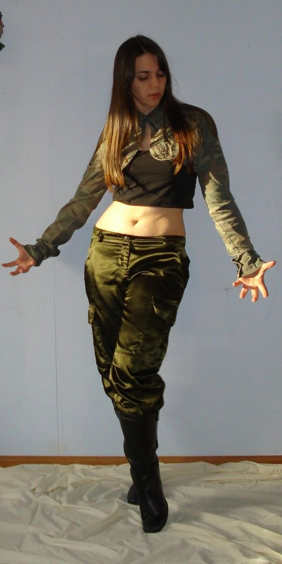 a young woman with long brown hair is standing in a room with soft, natural light. she is wearing olive green satin cargo pants, a black cropped top, and a camouflagepatterned jacket with threequarter sleeves. the jacket is open, revealing her midriff.  her black boots reach up to her midcalf. she is striking a pose with her arms outstretched, palms open. the background is a simple offwhite wall and the floor appears to be covered with a lightcolored fabric.
