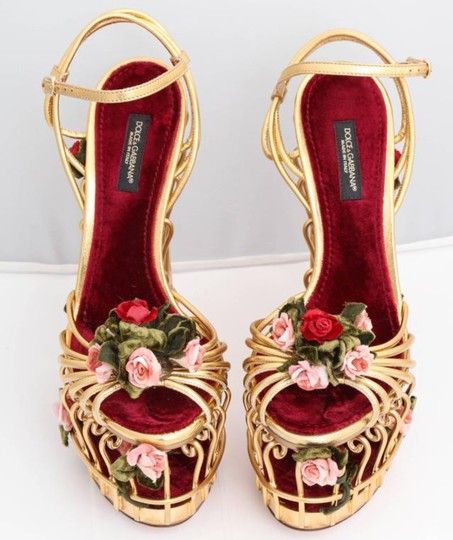the image showcases a pair of dolce & gabbana high heels.  these platform heels are a striking gold metallic color with a burgundy red velvet interior.  the shoes feature a cagelike design on the front, intricately decorated with delicate pink and red rose embellishments. the brand's logo is visible inside the shoes. they appear to be elegant and highfashion footwear suitable for a formal occasion or special event.