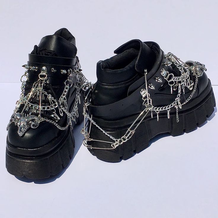 a pair of black, platform gothicstyle shoes heavily adorned with metal chains, spikes, safety pins, and other embellishments are positioned on a light, neutral surface. the shoes are leather and feature thick platform soles with a heavily textured grip. the shoe on the left is decorated with a buckle strap, studs, and an ornate metal charm in the shape of a goat's head. both shoes have chains dangling with small metallic pendants and safety pins connected to the platform.