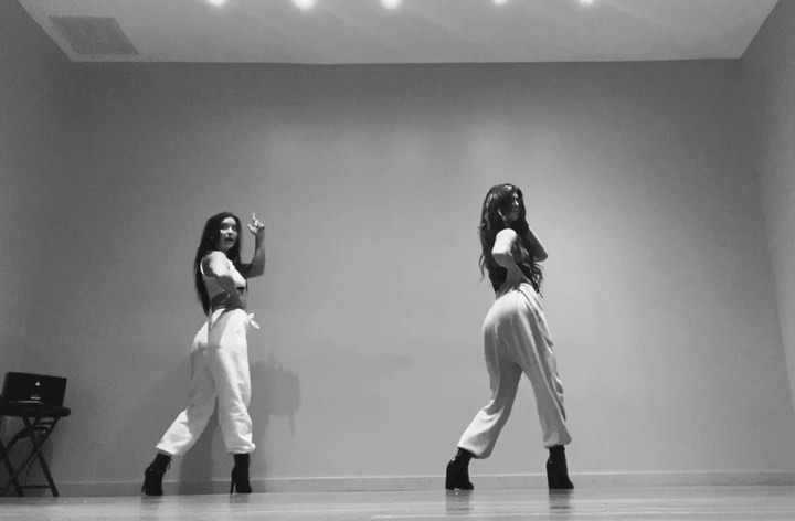 two women in white trousers and heels are dancing in a studio. the woman on the left is pointing up. the image is black and white.