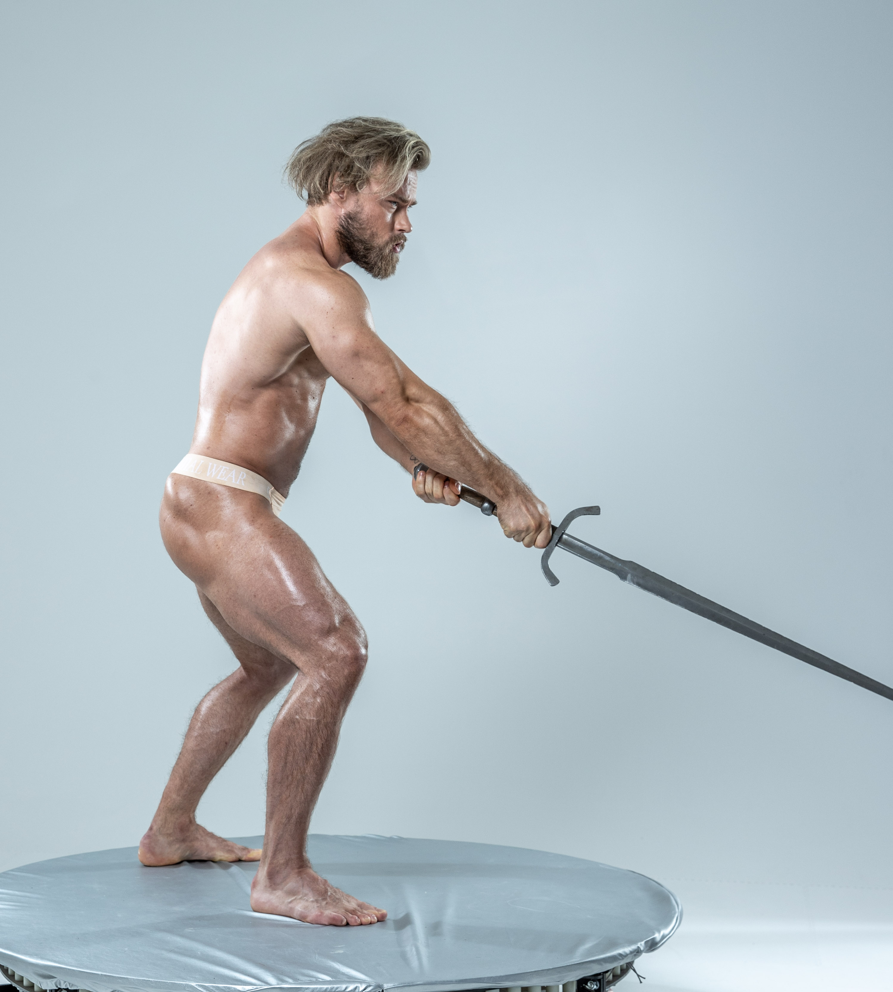 a muscular man with a chiseled physique is captured in a powerful pose against a gray background. his blonde hair is tousled, and he has a thick beard. he wears only a small, beige loincloth, which accentuates his welldefined muscles. in his hands, he grips a doubleedged sword, its polished metal glinting under the studio lights. he holds the sword with both hands, positioned as if about to strike or parry an attack. the man's focused expression and taut muscles convey a sense of controlled power and readiness for battle.  he stands on a circular platform, suggesting a staged environment, perhaps for photography or performance.
