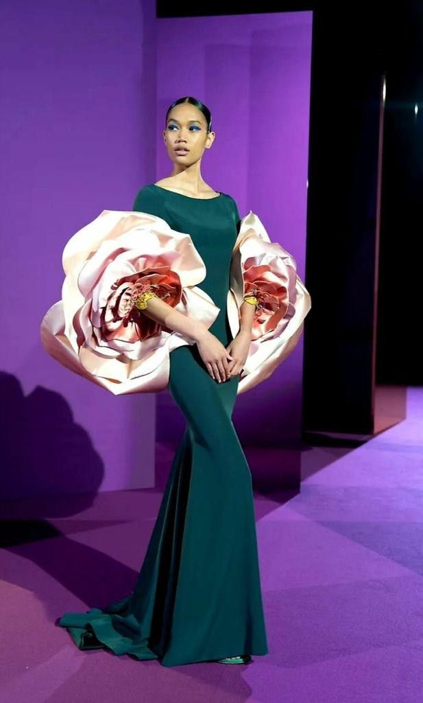 a female model in a green, floorlength dress with a large, ruffled floral design poses on a purple runway. the background is a blurred purple backdrop with a mirror reflecting the model. she has bright, bold makeup.