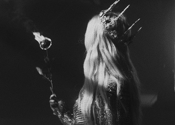 a black and white image shows a woman from the back, wearing a crown with prominent spikes or thorns. she has long hair flowing down her back and is holding a rose, the flower head appearing burnt or in flames. the overall atmosphere is dark and mysterious.