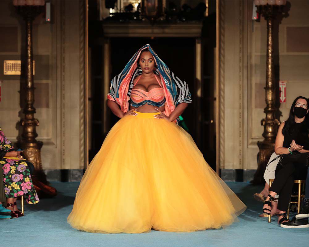 a black woman in a yellow tulle gown walks the runway during a fashion show. the gown has a black and white striped bodice and a pink and blue scarf. she is walking towards the camera and has a confident expression on her face. the audience is visible in the background.