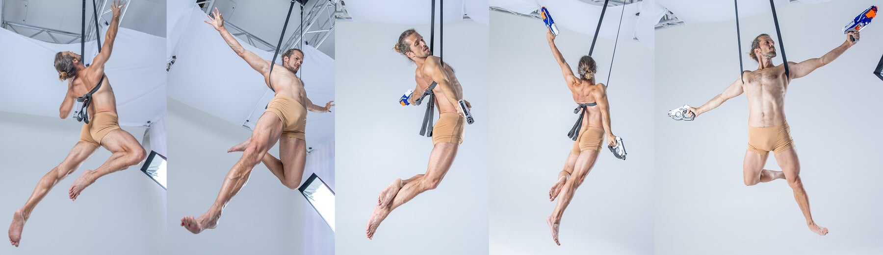 a collage of six images, all featuring the same shirtless muscular man. he is suspended from the ceiling by straps that are attached to a harness. he is in midair, and he is flexing his muscles as he stretches and jumps. he is holding a toy gun in a couple of the images, but in most he is just enjoying his poses. the images are all set against a white backdrop.