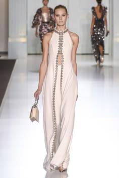 a  blonde haired model in a white floor length  gown,  with silver detailing is walking down the runway, during a high fashion show.