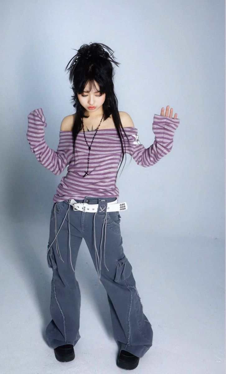 a full body shot of a young woman posing in a studio against a light blue background. she's wearing a purple and white striped offtheshoulder longsleeved shirt, grey cargo pants, a white belt with metal accents, and black shoes. her black hair is styled in a somewhat messy, choppy style, and she's wearing a simple cross necklace. her expression is downcast. the image showcases a grunge or alternative style.