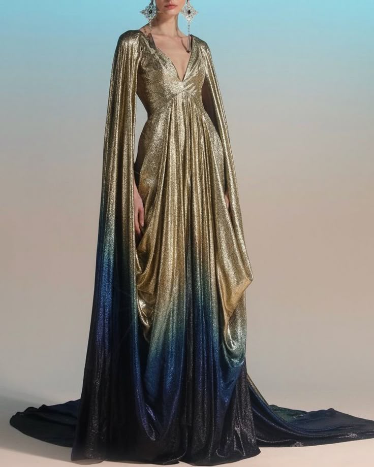 a woman is wearing a stunning, long, metallic gown. the dress is predominantly gold at the top, gradually transitioning into a blue and black ombre effect towards the bottom. it features a deep vneckline and draped details throughout the bodice and skirt. a dramatic cape, attached at the shoulders, extends down the length of the dress. the woman is also wearing statement earrings. the dress has a train, adding to its elegance and formality. the background is a gradient of blue and neutral tones.