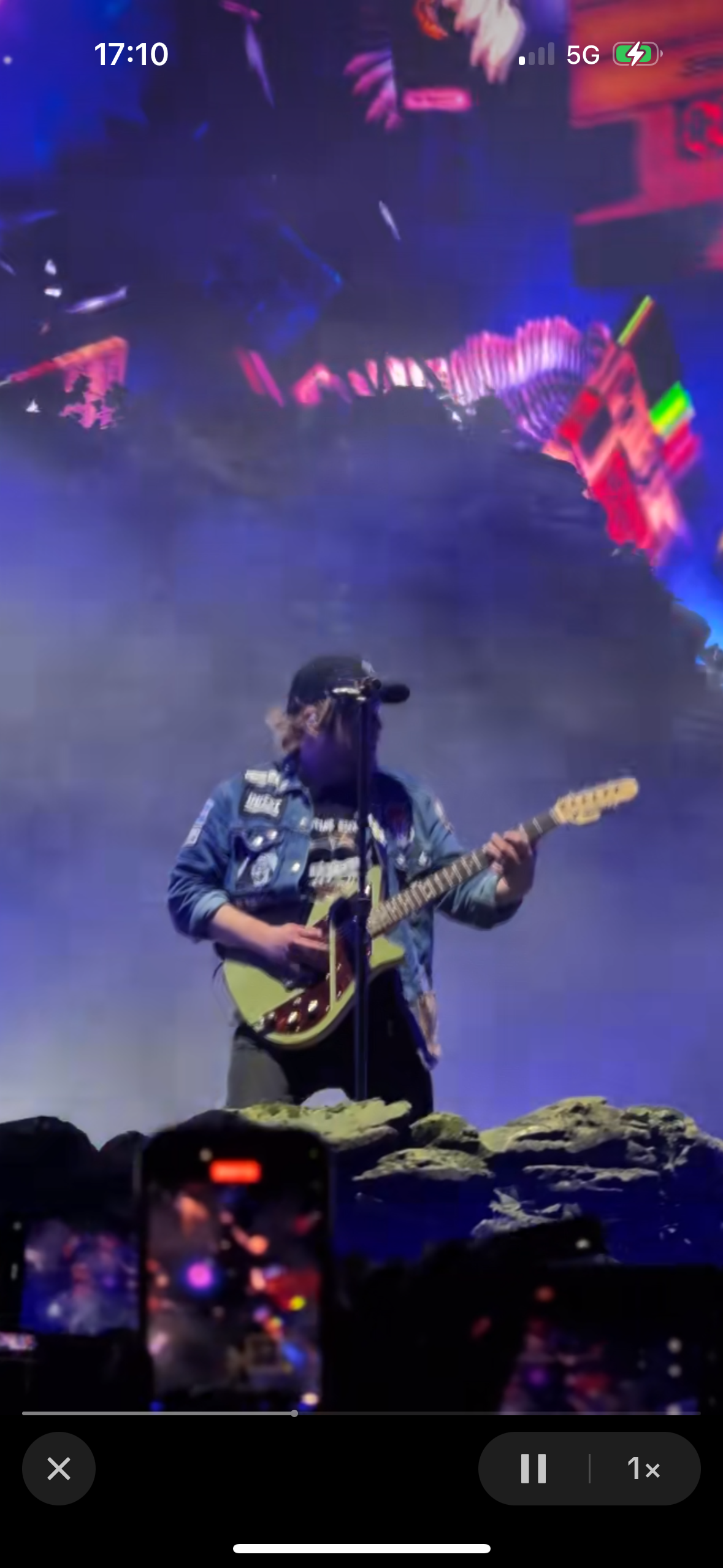a man plays electric guitar on stage during a concert. he is wearing a blue denim jacket and a black baseball cap, and he is holding his guitar with a passionate look on his face. the stage is lit up with colored lights and a crowd can be seen in the background. the image is full of energy and excitement.