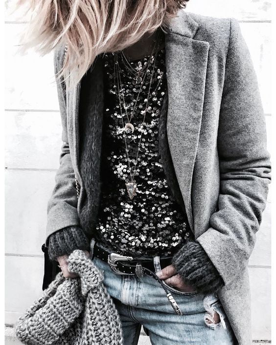 a woman is wearing a grey coat, a sparkly black top with a delicate silver necklace, and blue jeans with a ripped knee. she has a grey knit scarf draped over her shoulder and a silver belt with a large buckle. she is holding her grey scarf in her right hand and posing with a slightly sideward look. her blonde hair is loosely styled and framing her face.