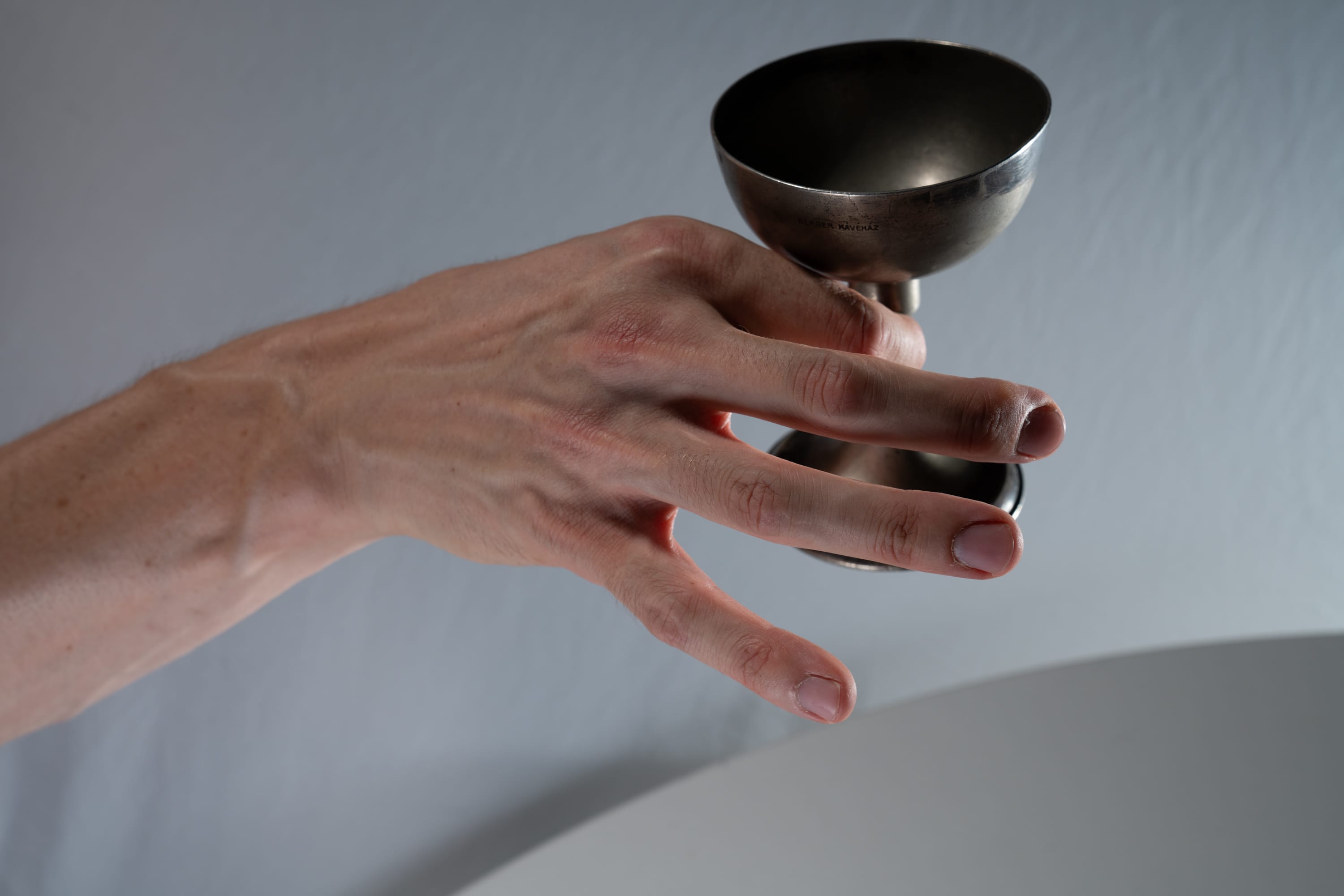 a closeup of a fairskinned hand delicately holding a small silver cup against a minimalist grey and white background. the soft lighting emphasizes the textures and details of the hand and the cup.