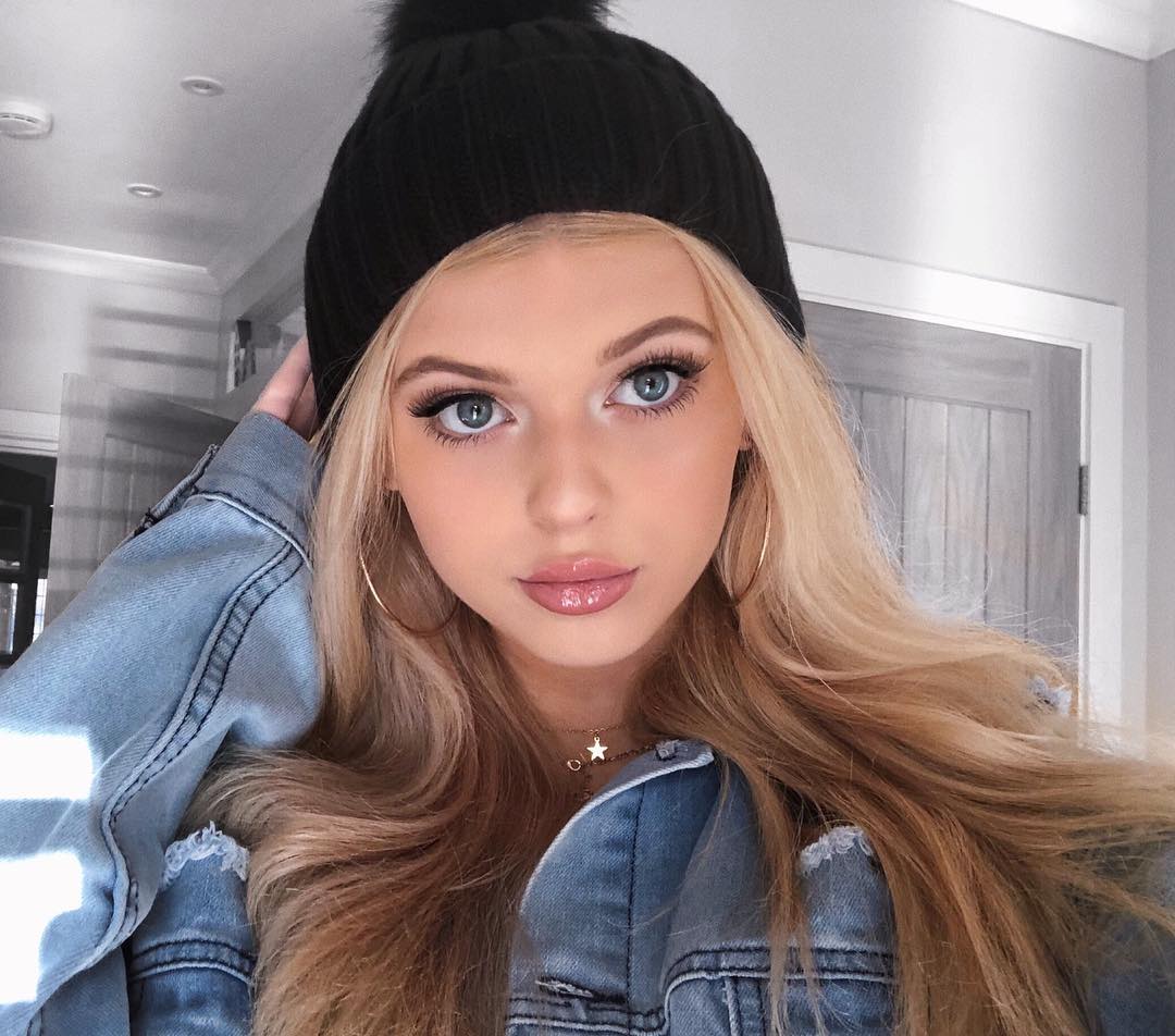 a closeup shot of a woman with blonde hair wearing a black beanie, a denim jacket, and gold hoop earrings. she has blue eyes and is wearing makeup, including lipstick. she is also wearing a necklace with a star pendant.
