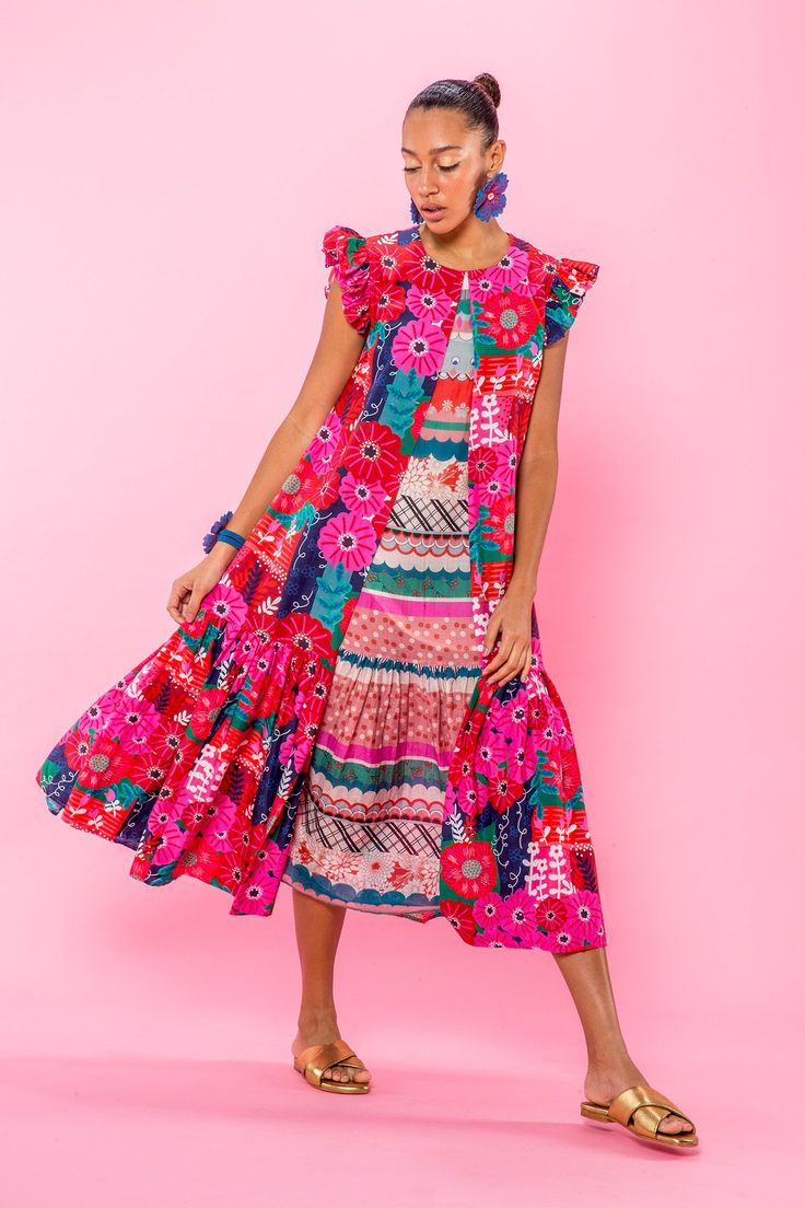 a fullbody shot of a woman wearing a vibrant, colorful midi dress with a floral print. the dress has ruffles and a sleeveless design.  the woman is posing against a solid pink background. she is wearing gold sandals and blue flower earrings. the overall style is bohemian and eclectic. the image is welllit and the colors are saturated.