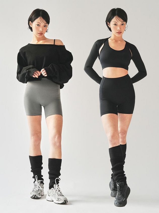 two women stand sidebyside against a white background modeling athletic wear. the woman on the left wears a black offtheshoulder sweater, gray shorts, black leg warmers, and gray and white sneakers. the woman on the right wears a black sports bra with a longsleeved shrug, black shorts, black leg warmers, and black sneakers. both women have short black hair.