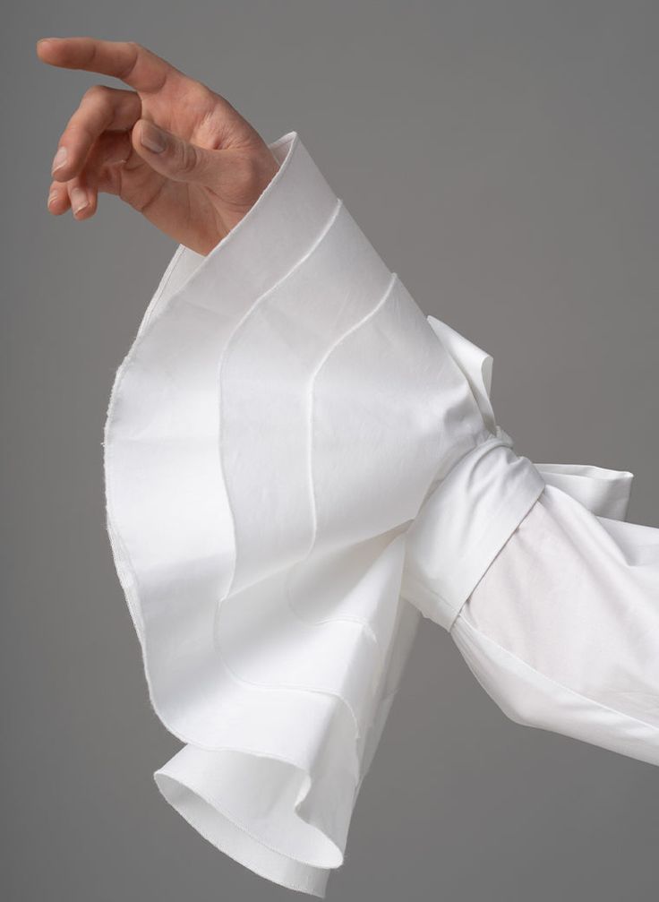a closeup shot of a woman's hand wearing a white shirt with a ruffled sleeve. the shirt is made of a delicate, textured fabric and the ruffle is very voluminous, adding an elegant touch to the garment.
