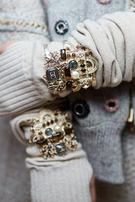 loseup shot of a person wearing a beige knit sweater and two elaborate chanel bracelets. the bracelets are large and ornate, featuring gold metalwork, various stones, pearls, and the chanel logo. the focus is on the intricate detail of the jewelry and its contrast against the soft knit fabric of the sweater.  the image evokes a sense of luxury and winter fashion.